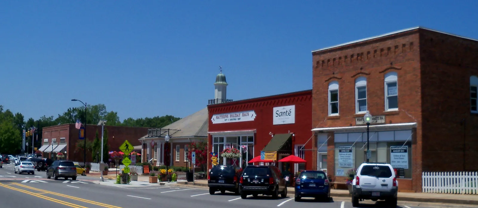 Brick storefronts along Trade Street in downtown Matthews, North Carolina, with red umbrellas, hanging flower baskets and a white picket fence under a clear blue sky