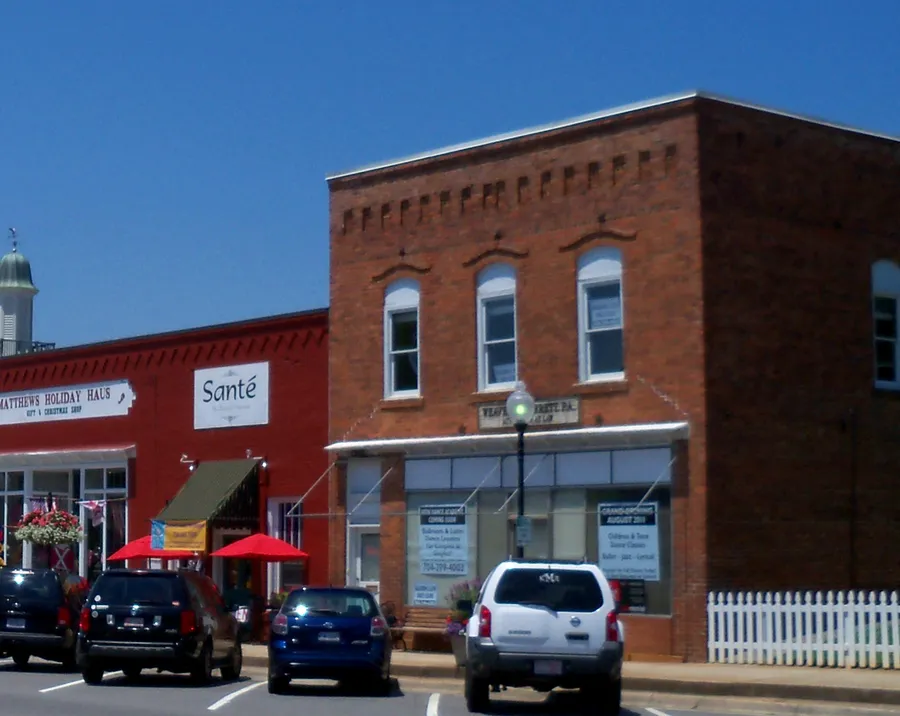 Brick storefronts along Trade Street in downtown Matthews, North Carolina, with red umbrellas, hanging flower baskets and a white picket fence under a clear blue sky